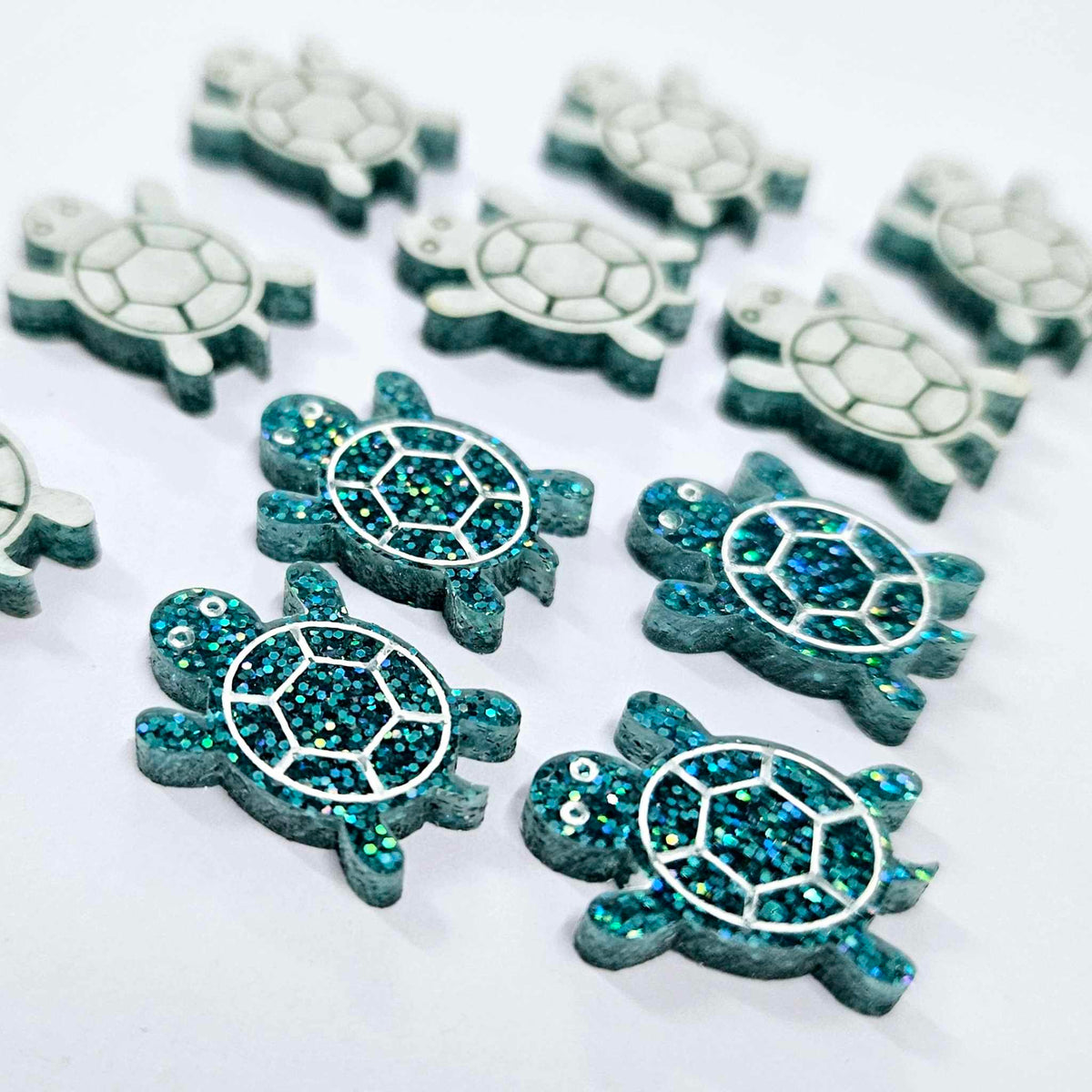 19mm teal glitter turtle acrylic stud earring blanks with engraved shell and eye details, some paint filled white, and some with protective paper still on.