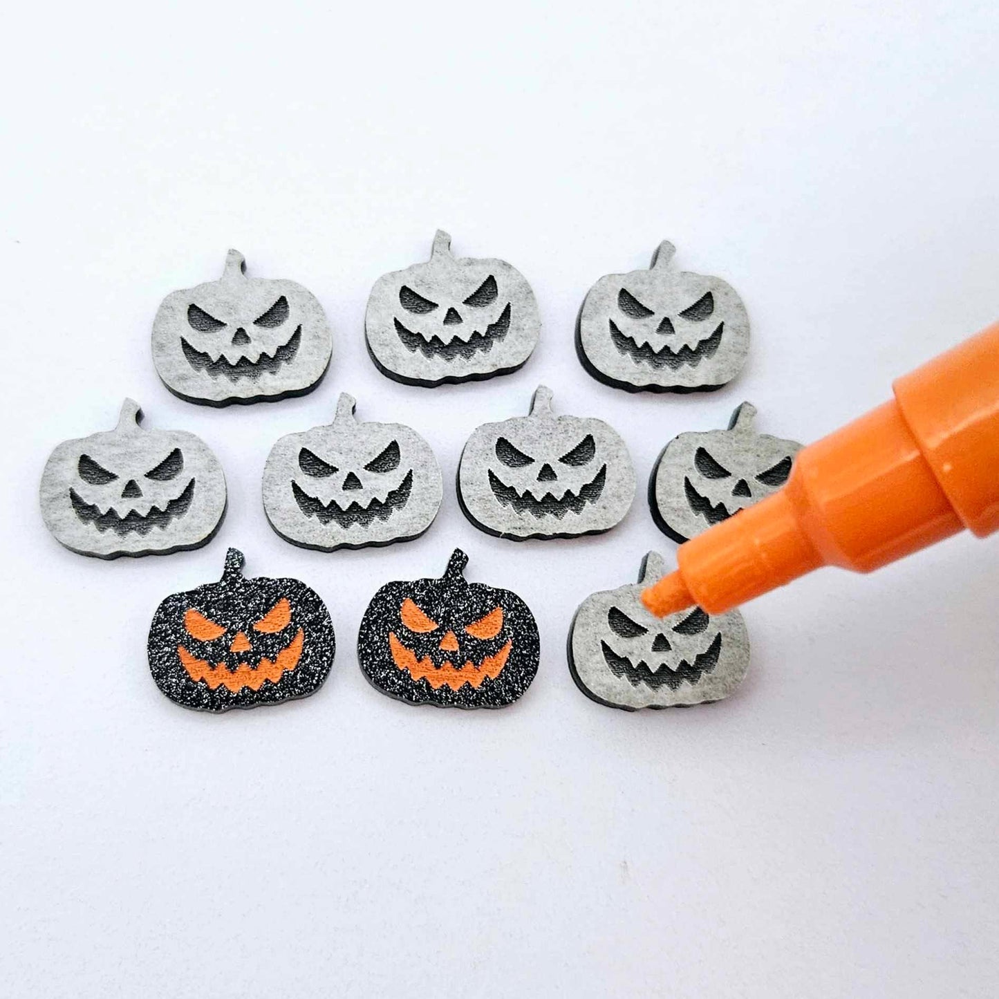 One pair of black glitter acrylic Jack-O’-Lantern stud earring blanks with orange paint-filled details, surrounded by multiple unpainted pieces with protective paper on, pictured with an uncapped Orange Posca Paint Pen ready for paint filling