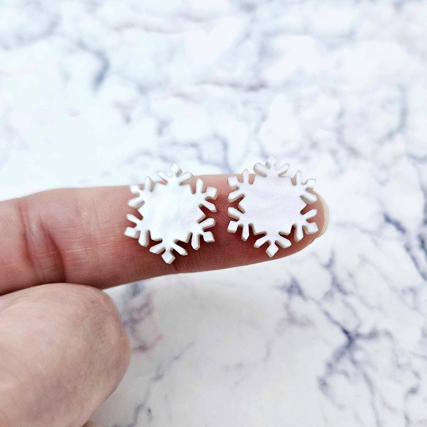One pair of 16mm white marble acrylic snowflake stud earring blanks shown on a finger for size reference.