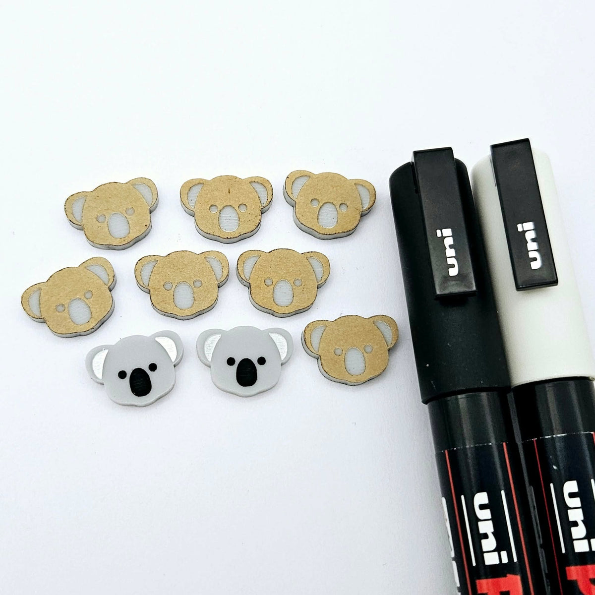 Solid Grey acrylic koala stud blanks showing one finished pair with protective paper removed and engraved details paint filled in black and white, alongside unpeeled blanks with protective paper still attached and POSCA paint pens used for decorating.