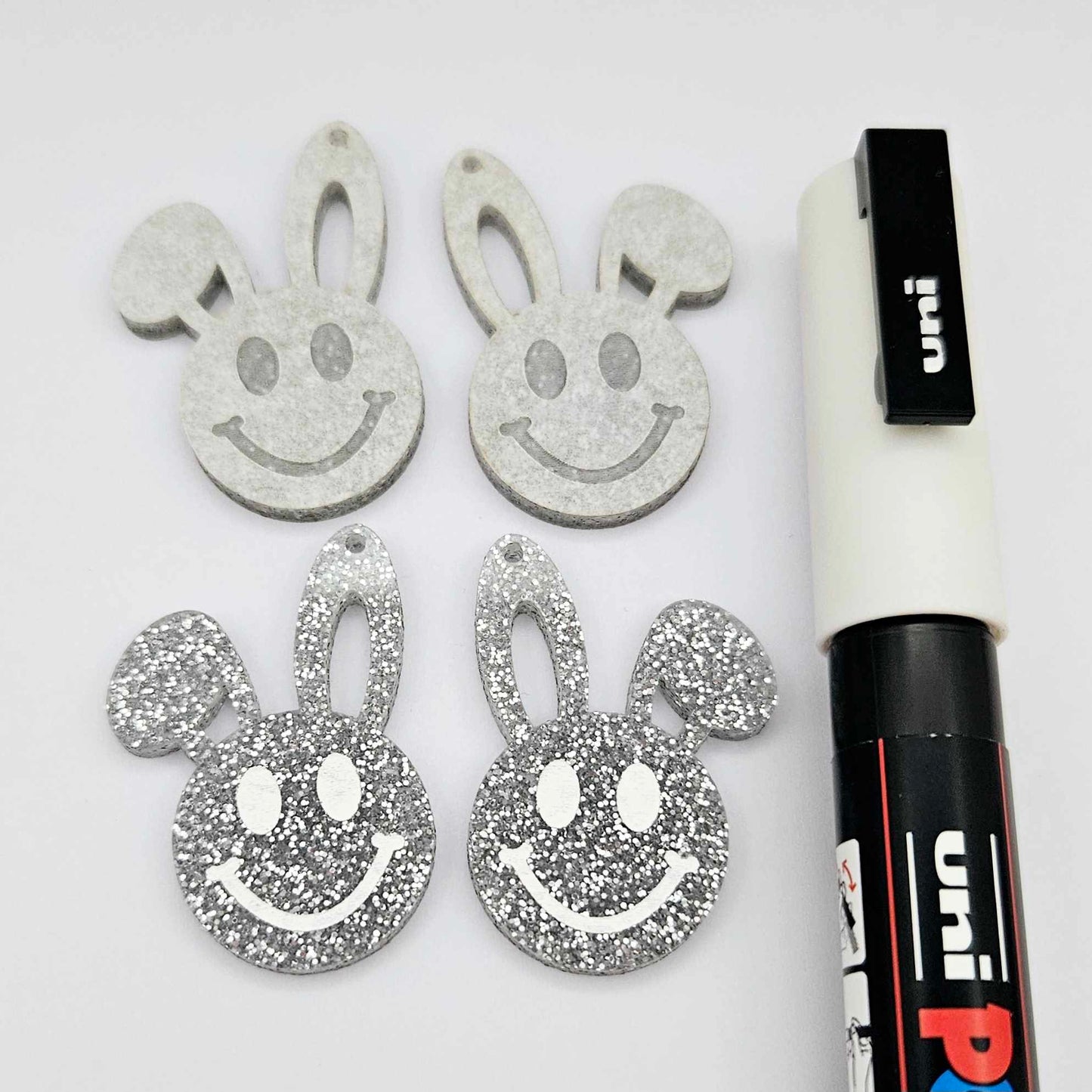 38mm silver glitter acrylic bunny dangle blanks with engraved smiley face, two paint filled white and two others unfilled with protective film on, shown with a White Posca paint pen