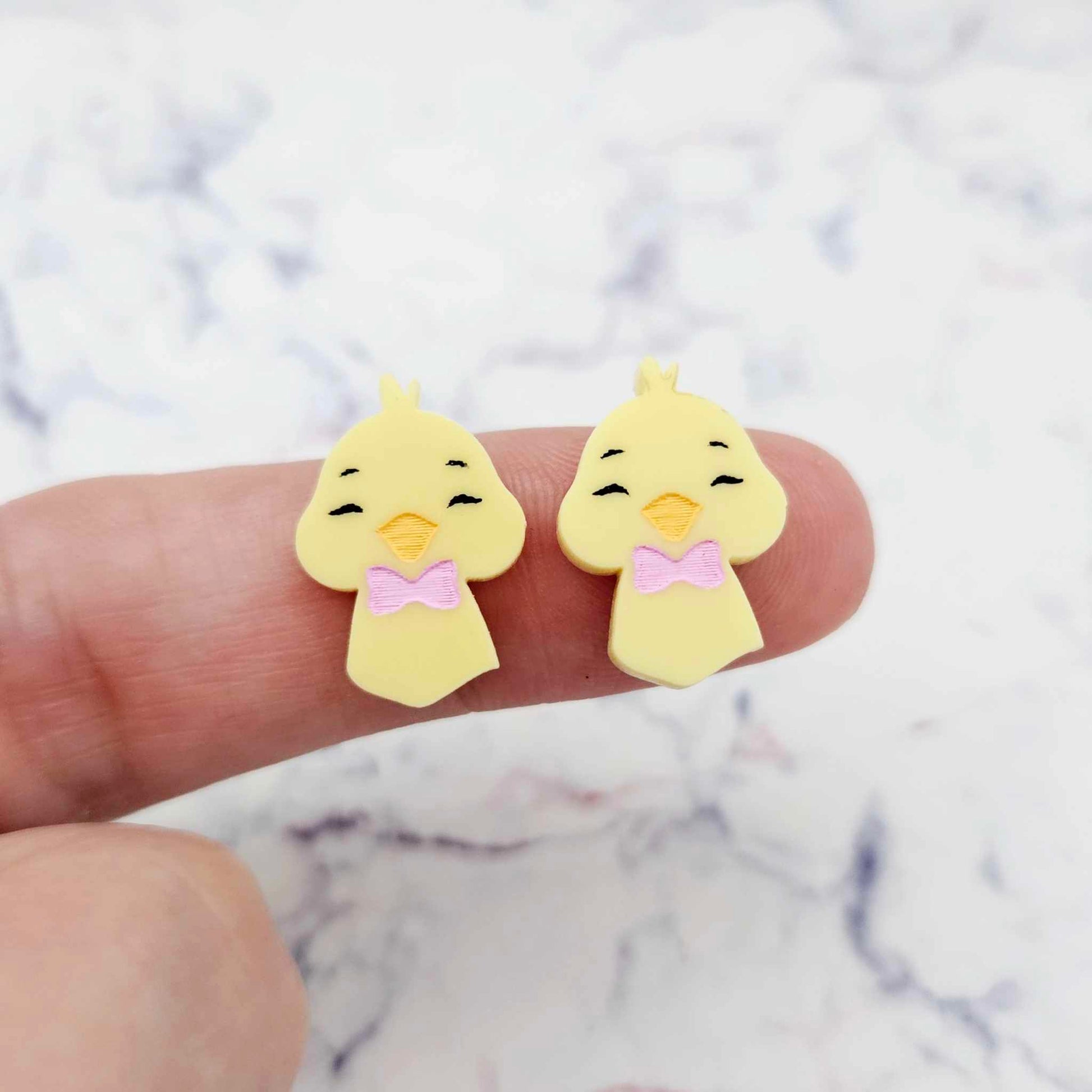 Close-up of two pastel yellow acrylic chicken head stud blanks with engraved face and bow tie, paint filled black, light pink and light orange