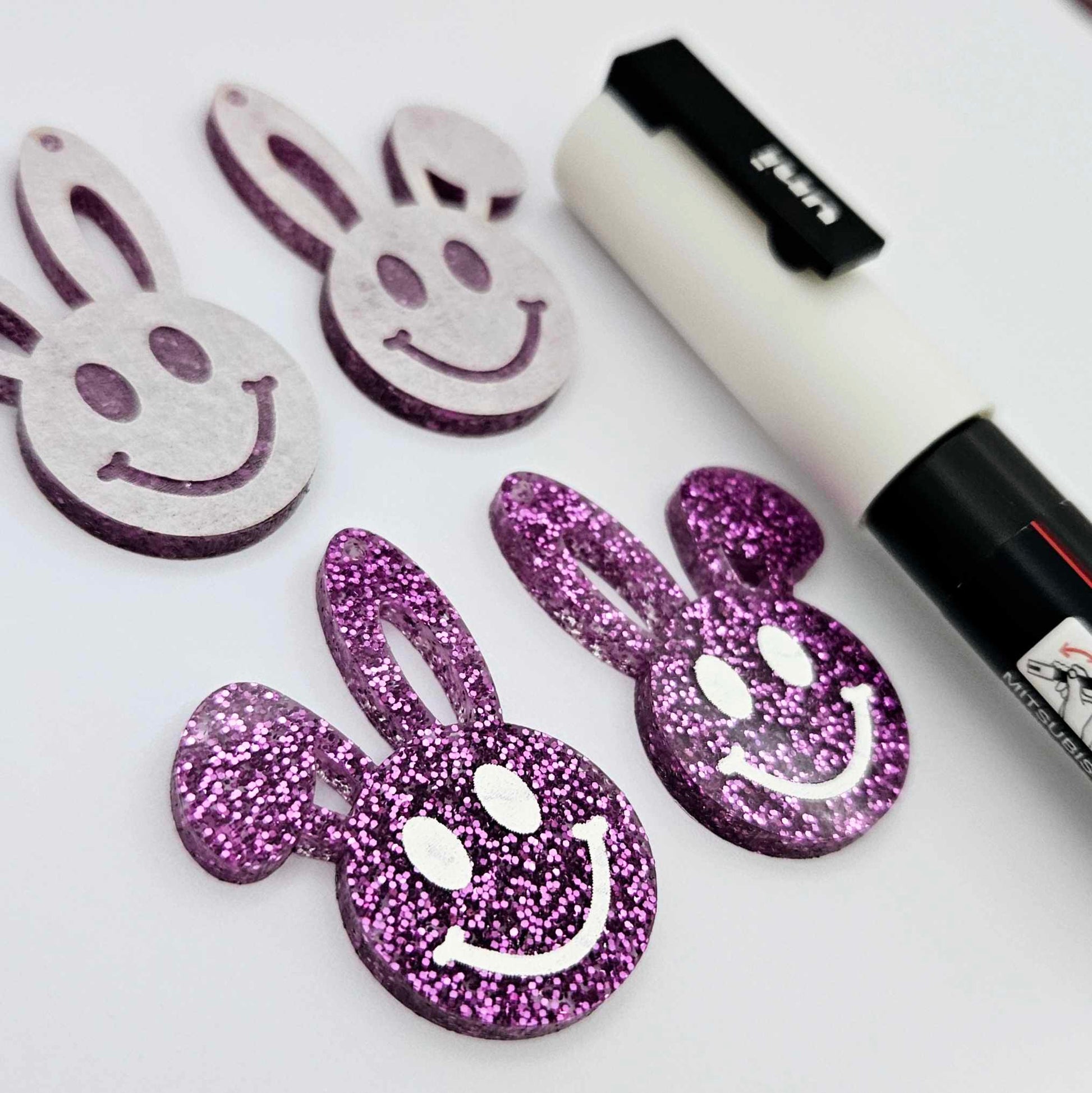 38mm purple glitter acrylic bunny dangle blanks with engraved smiley face, two paint filled white and two others unfilled with protective film on, shown with a White Posca paint pen