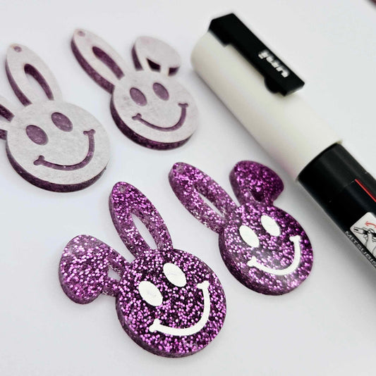38mm purple glitter acrylic bunny dangle blanks with engraved smiley face, two paint filled white and two others unfilled with protective film on, shown with a White Posca paint pen