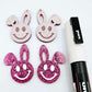 Hot pink glitter acrylic bunny dangle blanks with engraved smiley face, two paint filled white and two others unfilled with protective film on, shown with a White Posca paint pen