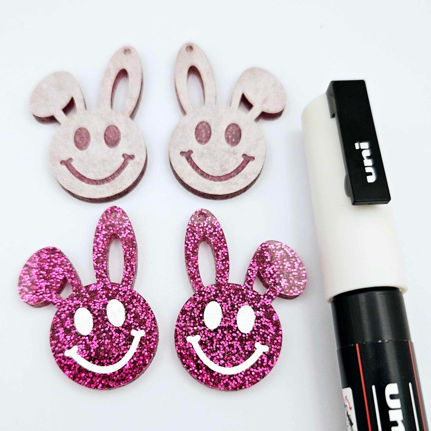 Hot pink glitter acrylic bunny dangle blanks with engraved smiley face, two paint filled white and two others unfilled with protective film on, shown with a White Posca paint pen