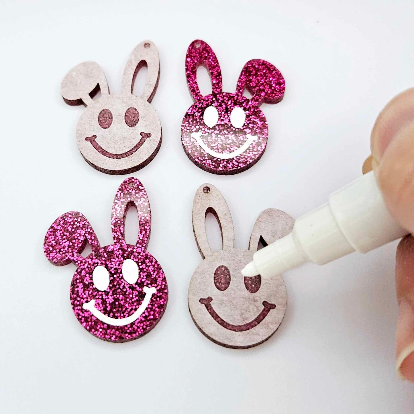 Hot pink glitter acrylic bunny dangle blanks with engraved smiley face, two paint filled white and two others unfilled with protective film on, shown with a White Posca paint pen