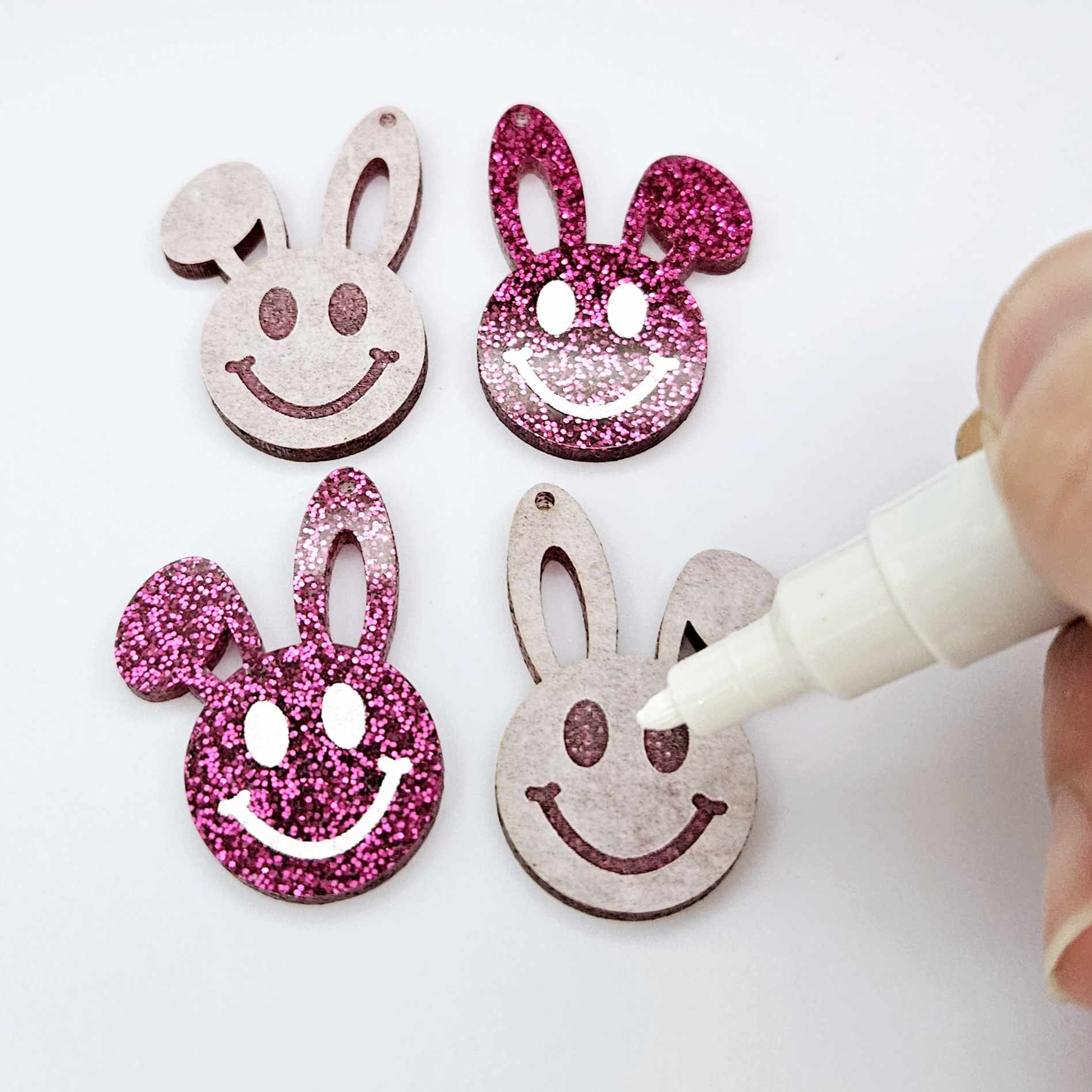 Hot pink glitter acrylic bunny dangle blanks with engraved smiley face, two paint filled white and two others unfilled with protective film on, shown with a White Posca paint pen