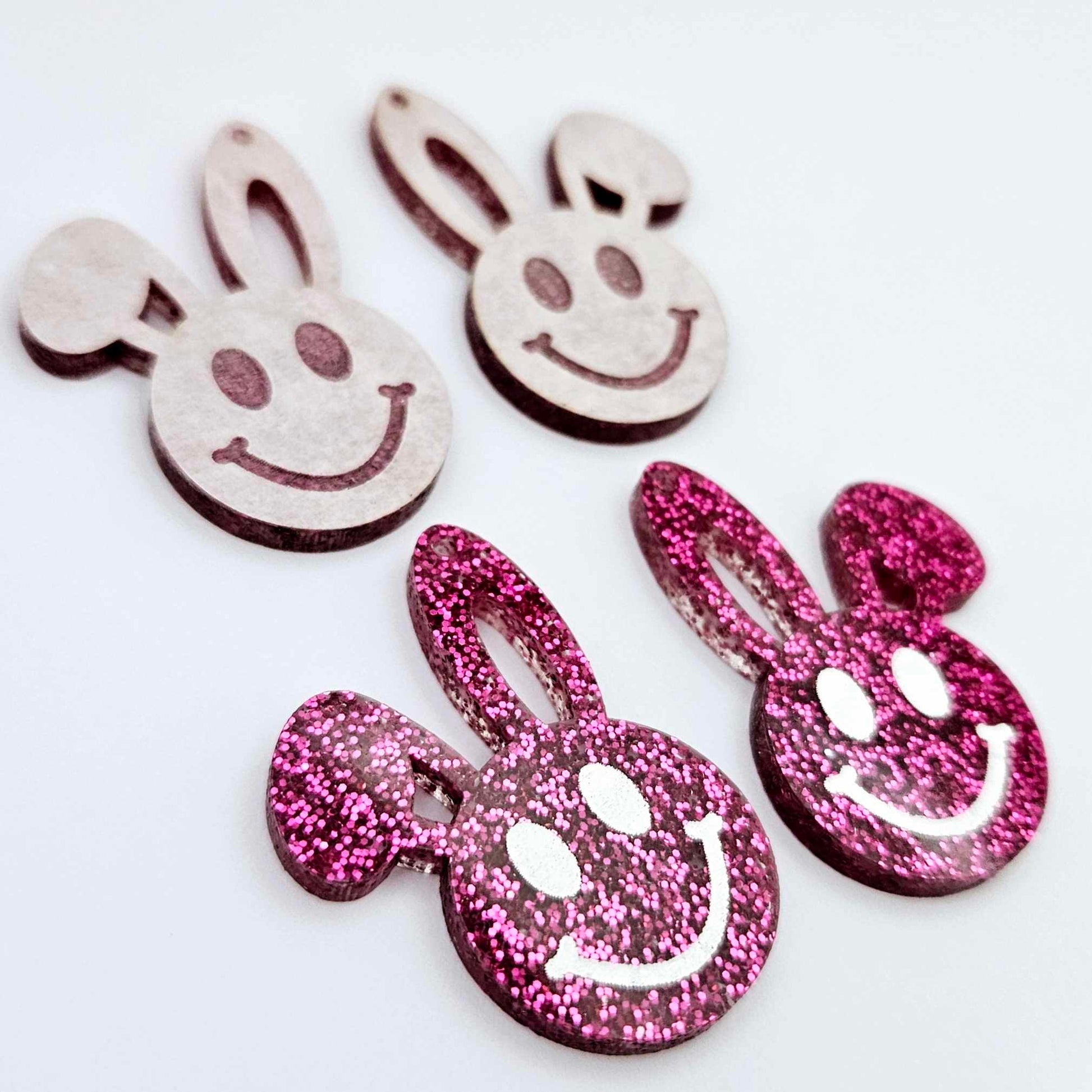 Hot pink glitter acrylic bunny dangle blanks with engraved smiley face, two paint filled white and two others unfilled with protective film on, shown with a White Posca paint pen