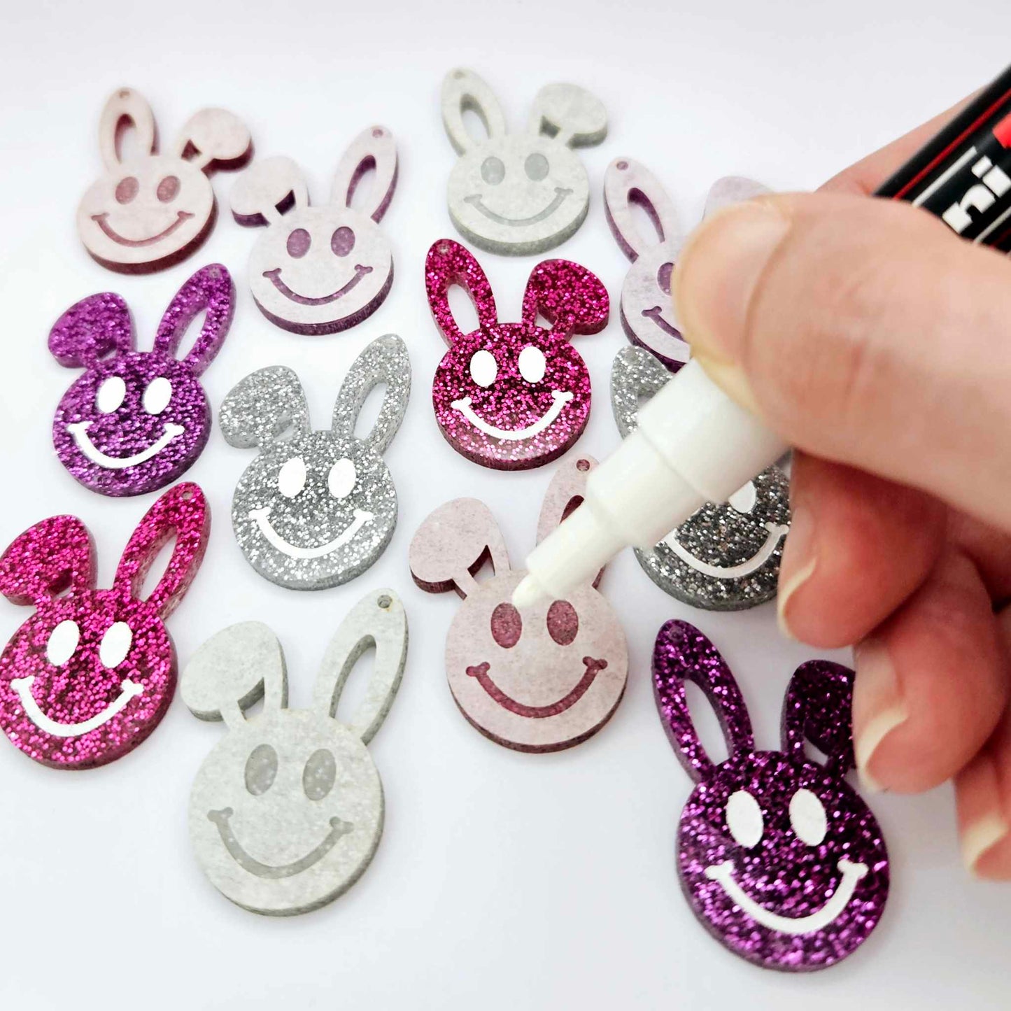 38mm silver, hot pink and purple glitter acrylic bunny dangle blanks with engraved smiley face, some paint filled white and others unfilled with protective film on, shown with a White Posca paint pen