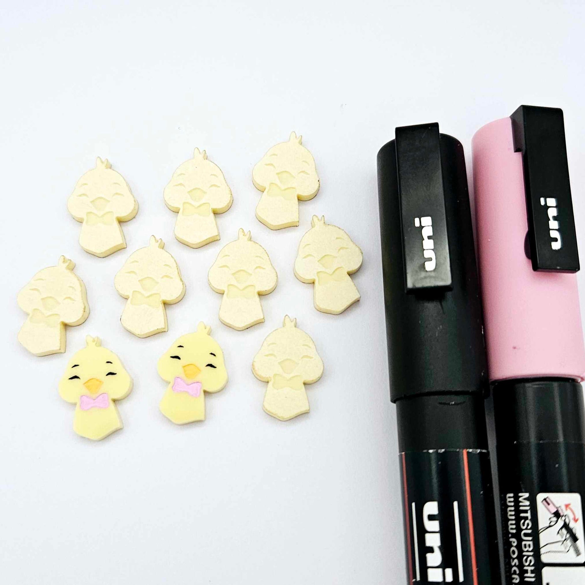 Pastel yellow acrylic chicken head stud blanks showing two paint filled examples and several unpainted pieces, with Posca paint pens included in the image