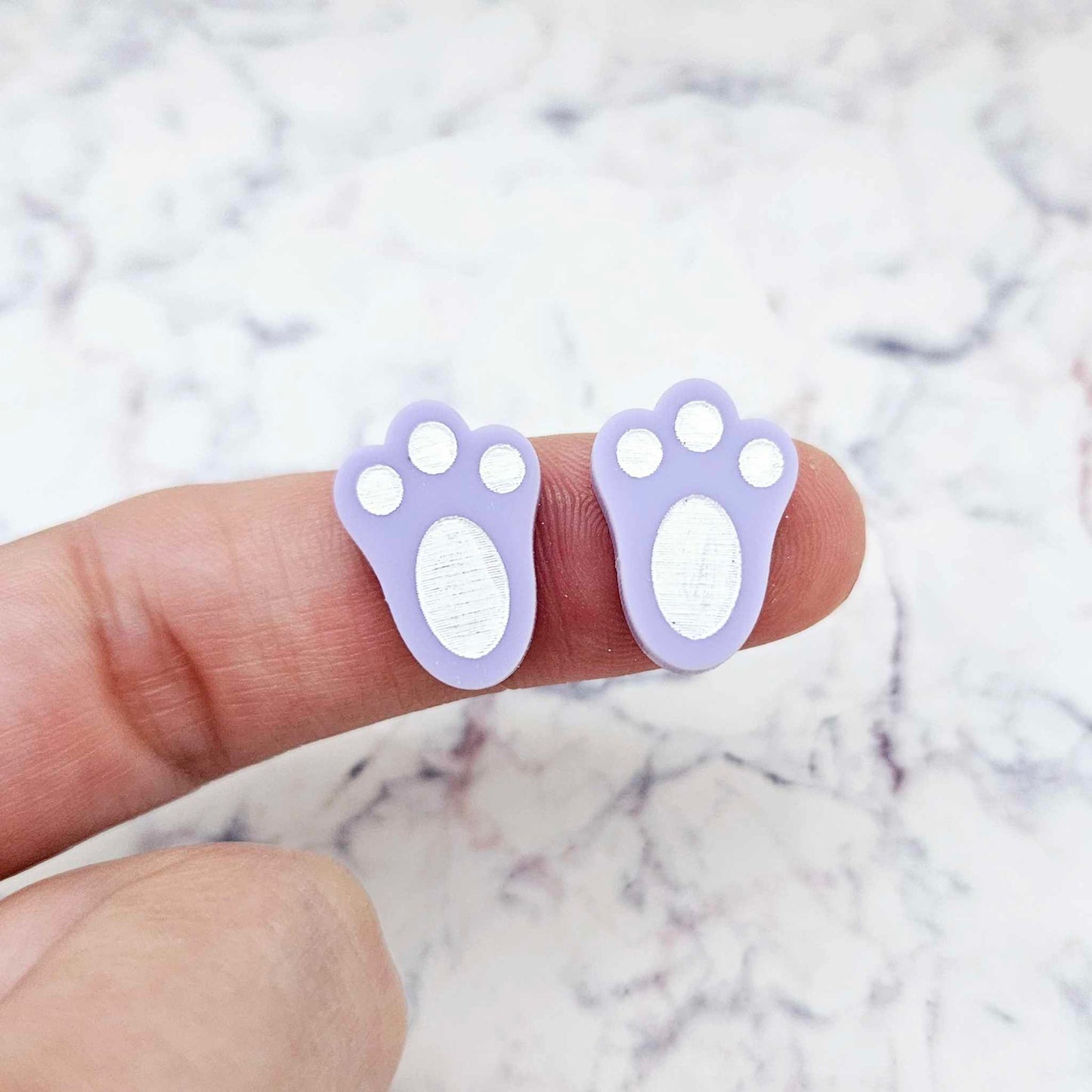 Close-up of two pastel purple acrylic bunny paw print stud blanks with engraved pads paint filled white