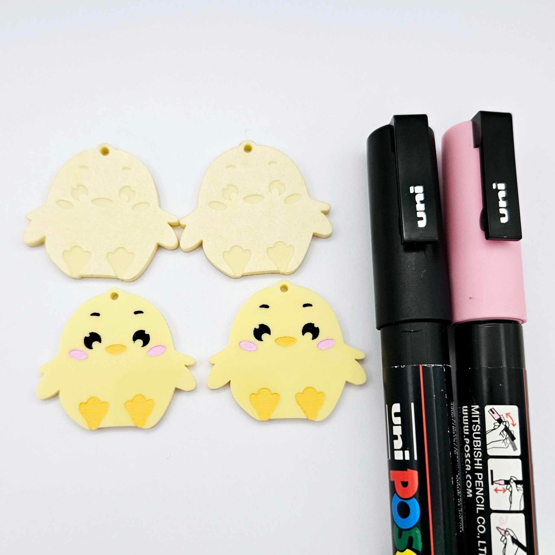 Pastel yellow acrylic chicken dangle blanks with engraved face and feet, two paint filled and two with protective film on, shown with Posca paint pens