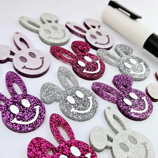 38mm silver, hot pink and purple glitter acrylic bunny dangle blanks with engraved smiley face, some paint filled white and others unfilled with protective film on, shown with a White Posca paint pen