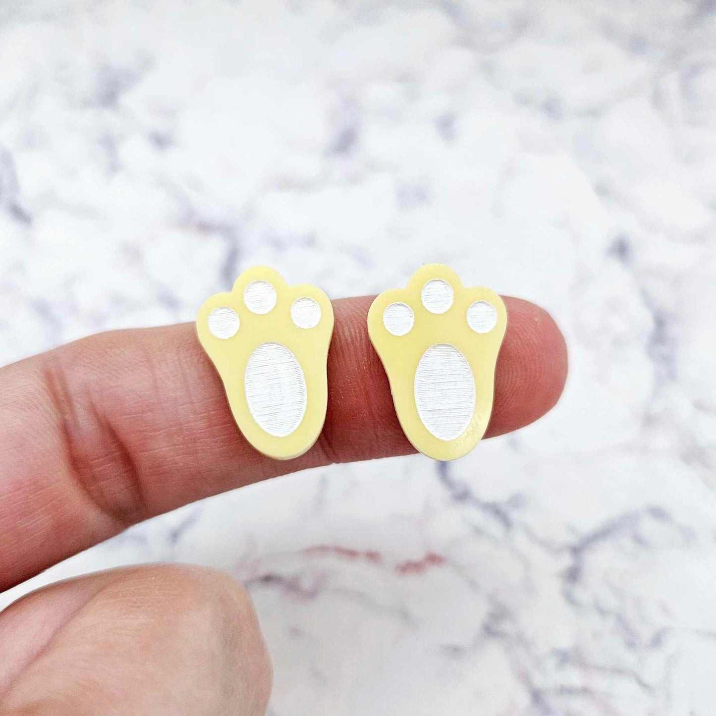 Close-up of two pastel yellow acrylic bunny paw print stud blanks with engraved pads paint filled white