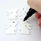 32mm white bunny dangle blanks showing paint filled black eyelashes on two pieces, with remaining pieces shown with protective paper as supplied