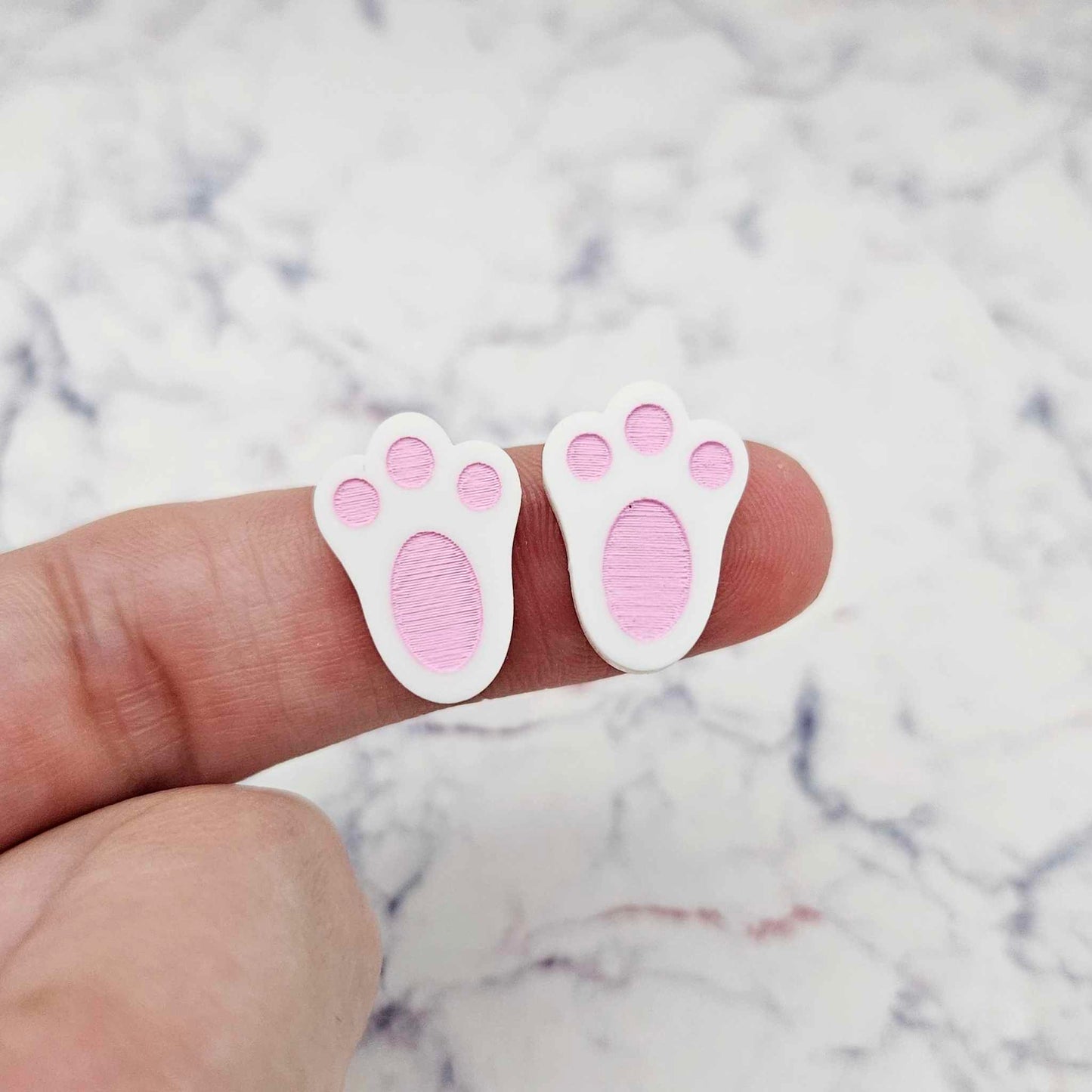 Close-up of two white acrylic bunny paw print stud blanks with engraved pads paint filled pink