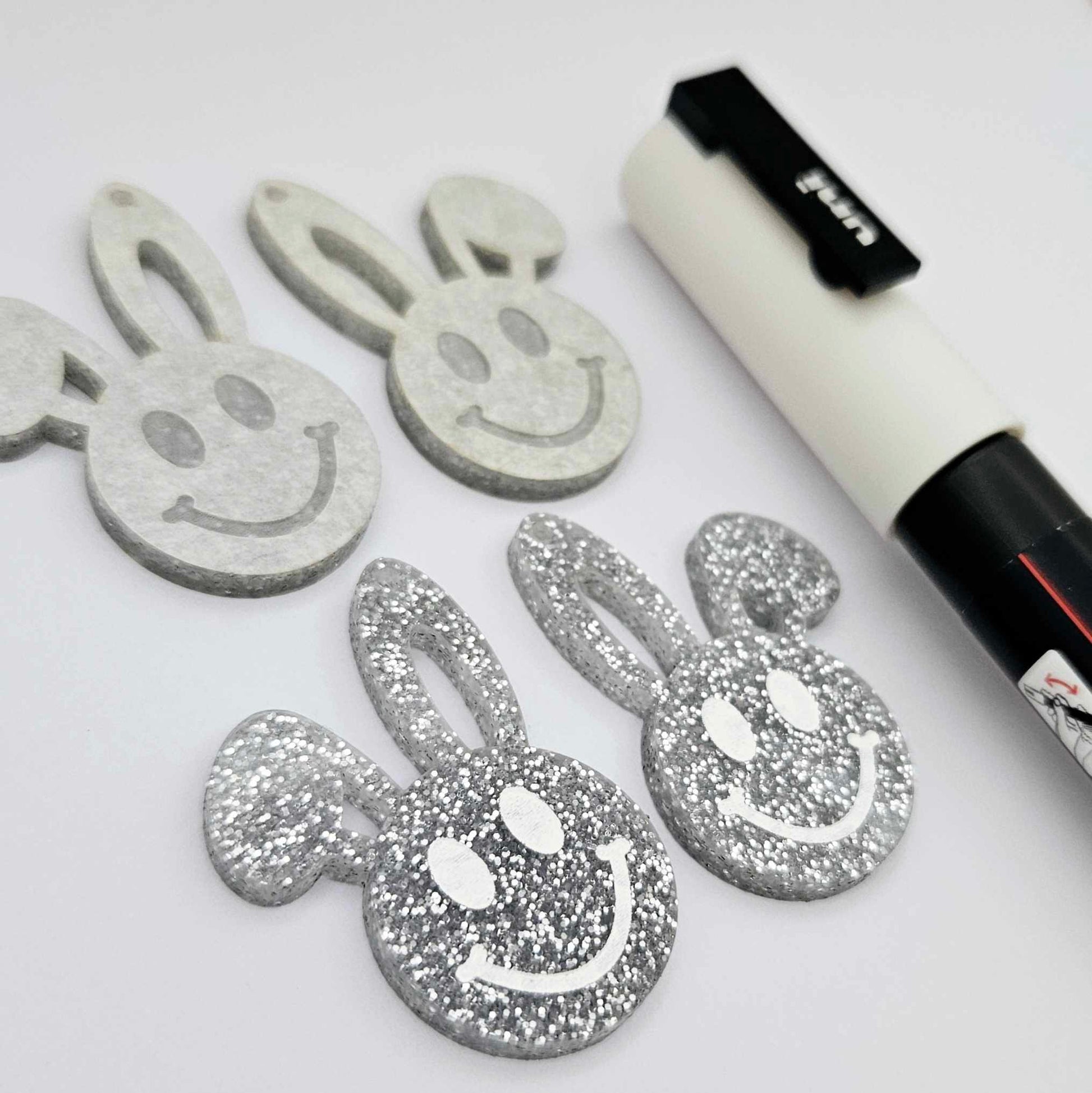 38mm silver glitter acrylic bunny dangle blanks with engraved smiley face, two paint filled white and two others unfilled with protective film on, shown with a White Posca paint pen