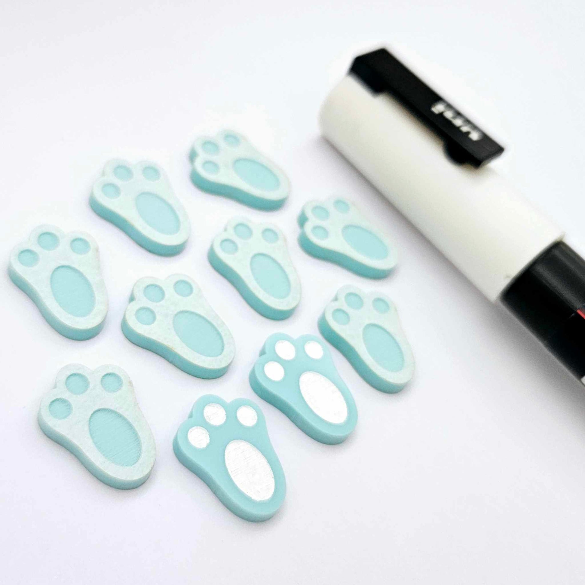 Pastel aqua bunny paw print stud blanks showing two paint filled white and several unpainted pieces, with a white Posca paint pen included in the image