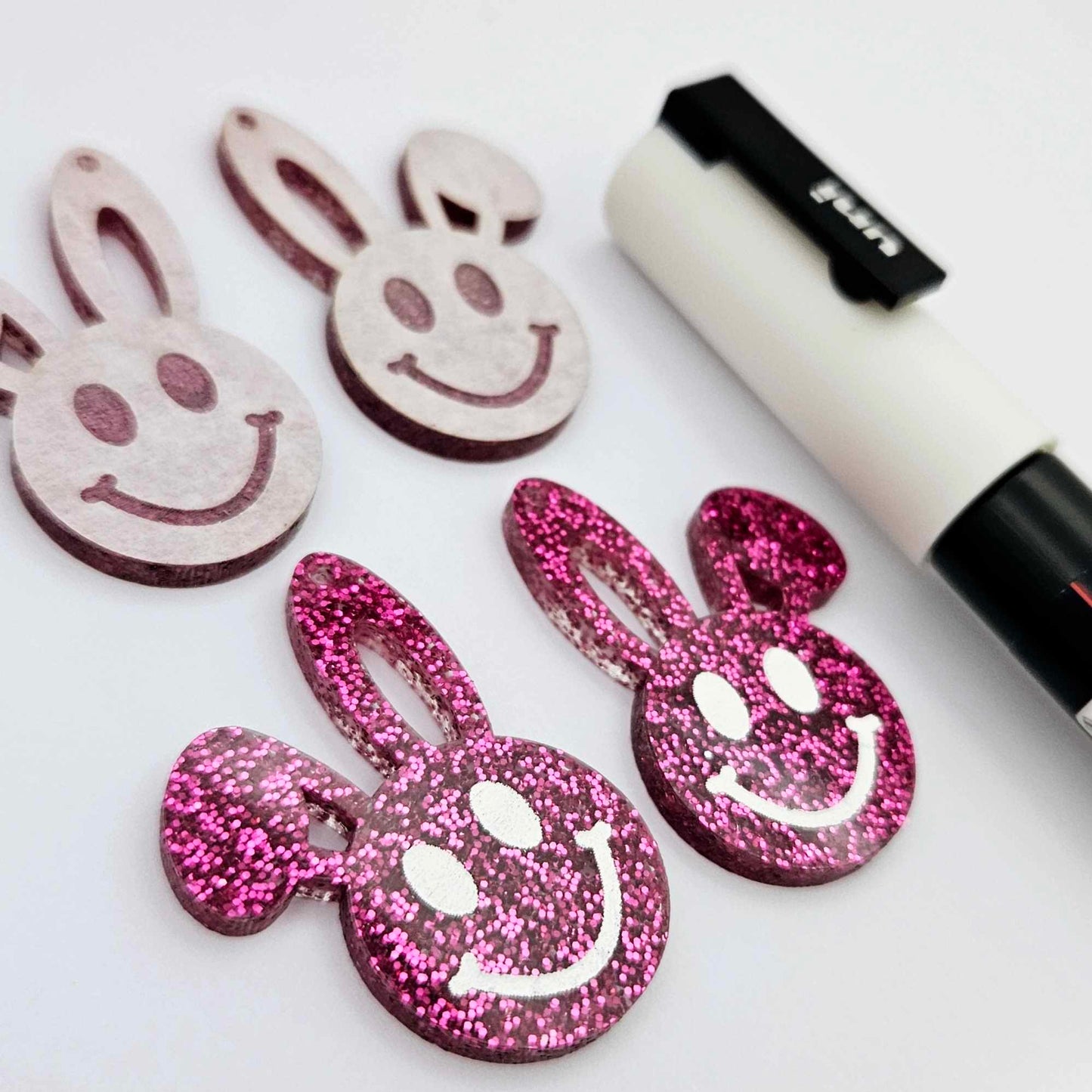 Hot pink glitter acrylic bunny dangle blanks with engraved smiley face, two paint filled white and two others unfilled with protective film on, shown with a White Posca paint pen