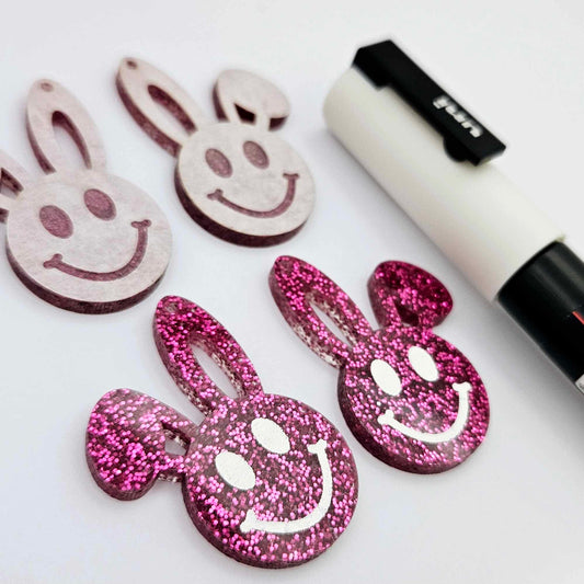 Hot pink glitter acrylic bunny dangle blanks with engraved smiley face, two paint filled white and two others unfilled with protective film on, shown with a White Posca paint pen