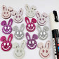 38mm silver, hot pink and purple glitter acrylic bunny dangle blanks with engraved smiley face, some paint filled white and others unfilled with protective film on, shown with a White Posca paint pen