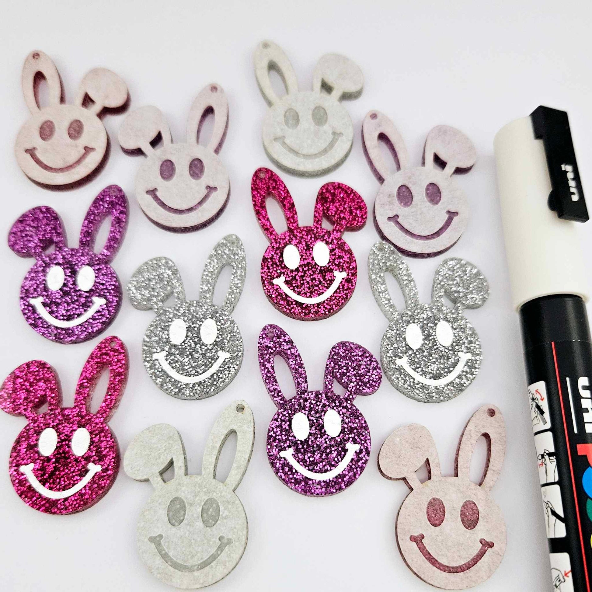 38mm silver, hot pink and purple glitter acrylic bunny dangle blanks with engraved smiley face, some paint filled white and others unfilled with protective film on, shown with a White Posca paint pen