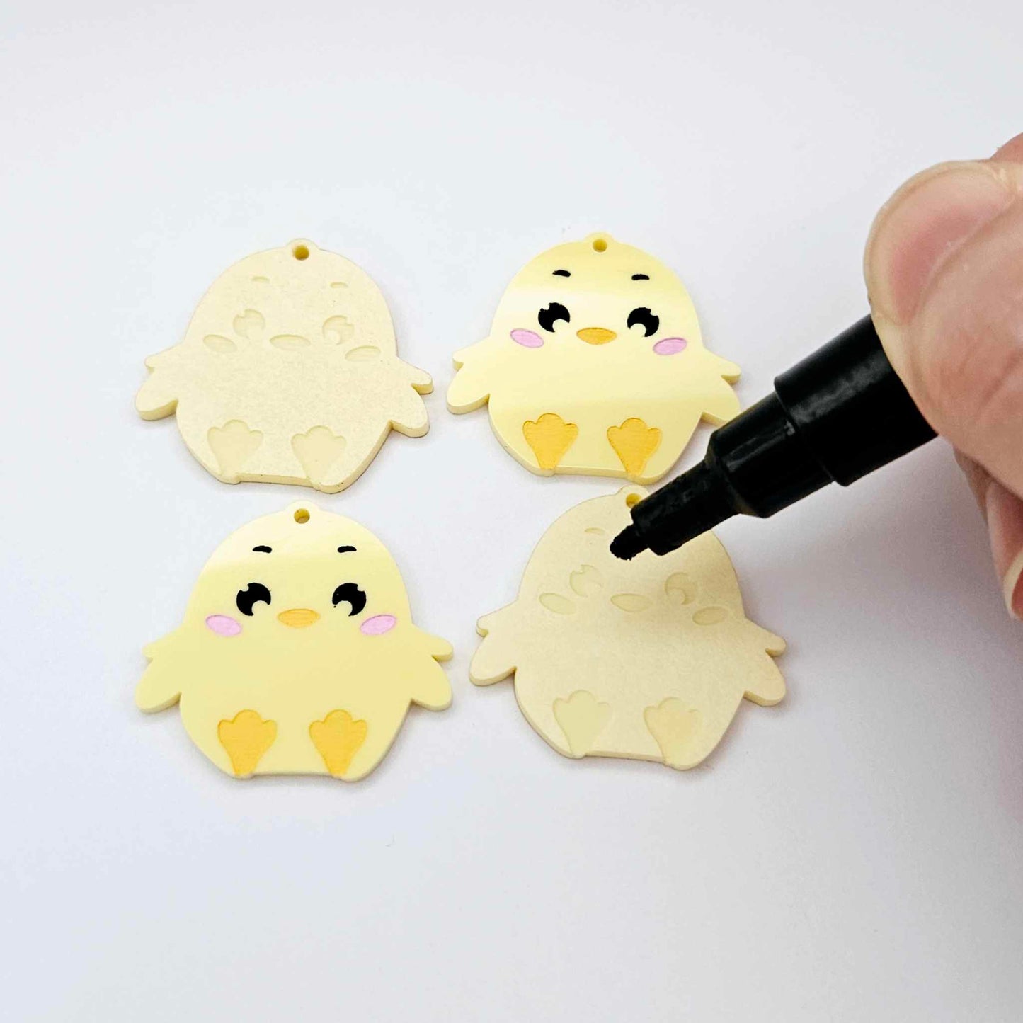 Pastel yellow acrylic chicken dangle blanks with engraved face and feet, two paint filled and two with protective film on, shown with a Posca paint pen