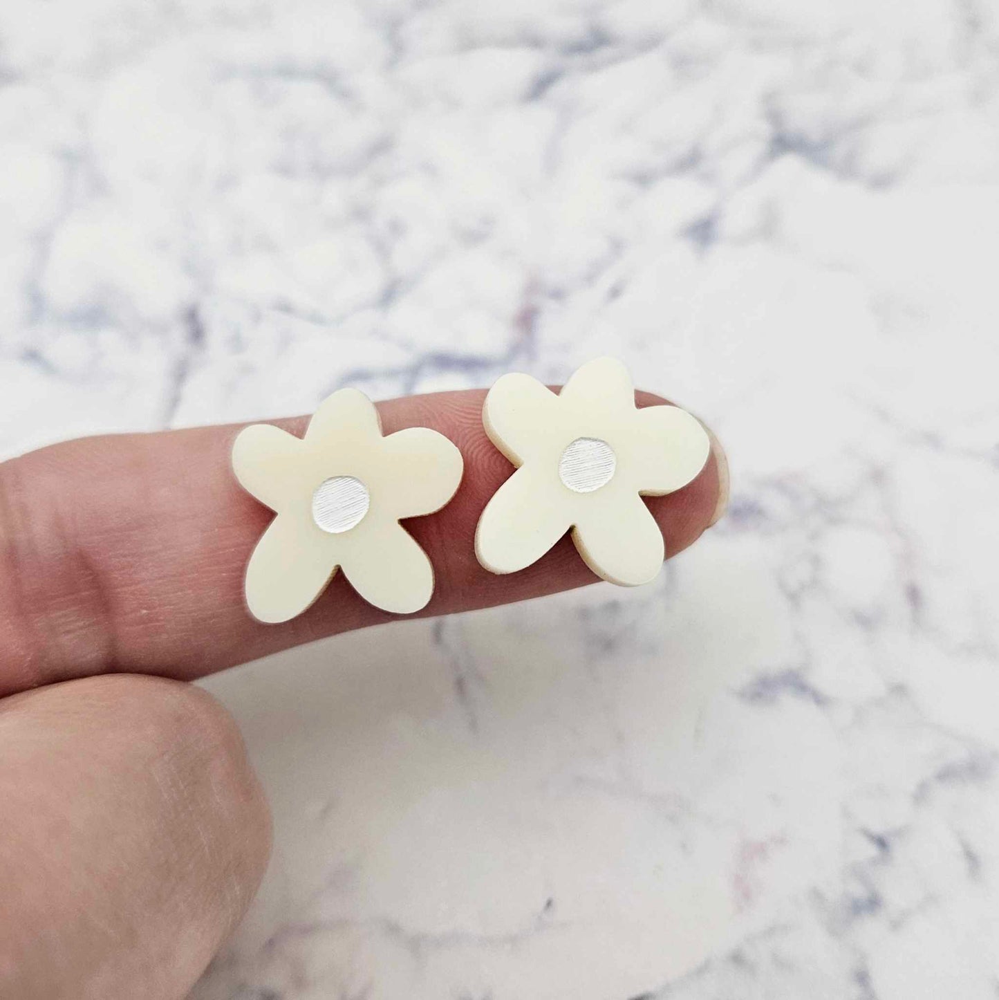 Two 16mm cream organic flower stud blanks with engraved centre paint filled white, shown on a finger for size reference