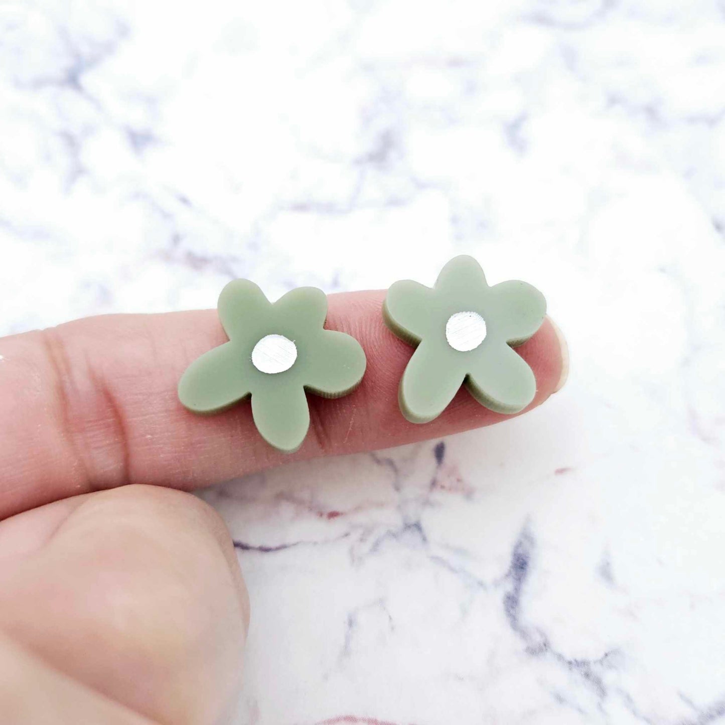 Two 16mm olive green organic flower stud blanks with engraved centre paint filled white, shown on a finger for size reference