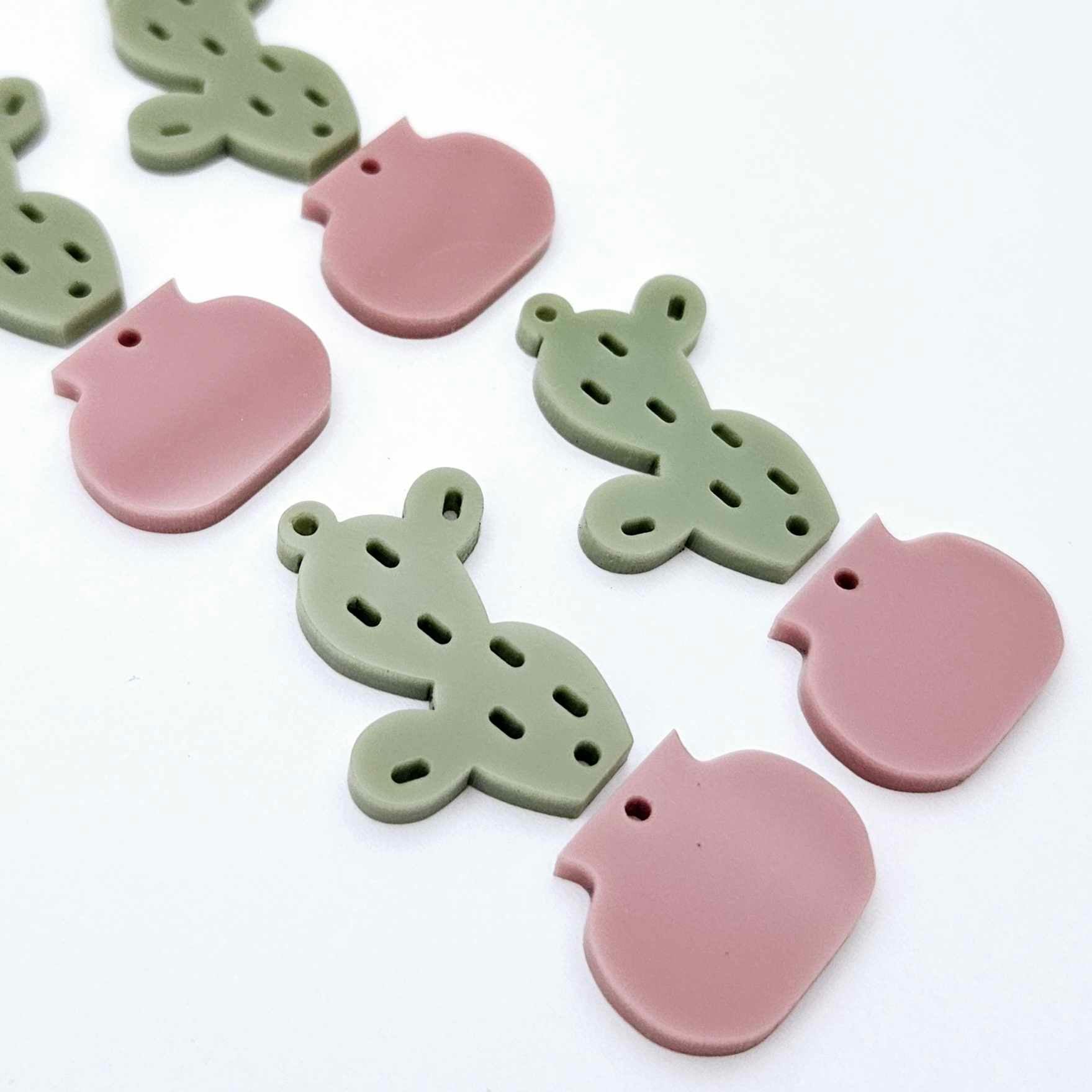 Two-piece acrylic cactus dangle blanks in olive green and dusty rose pink, shown unassembled with cactus top and pot bottom pieces laid flat