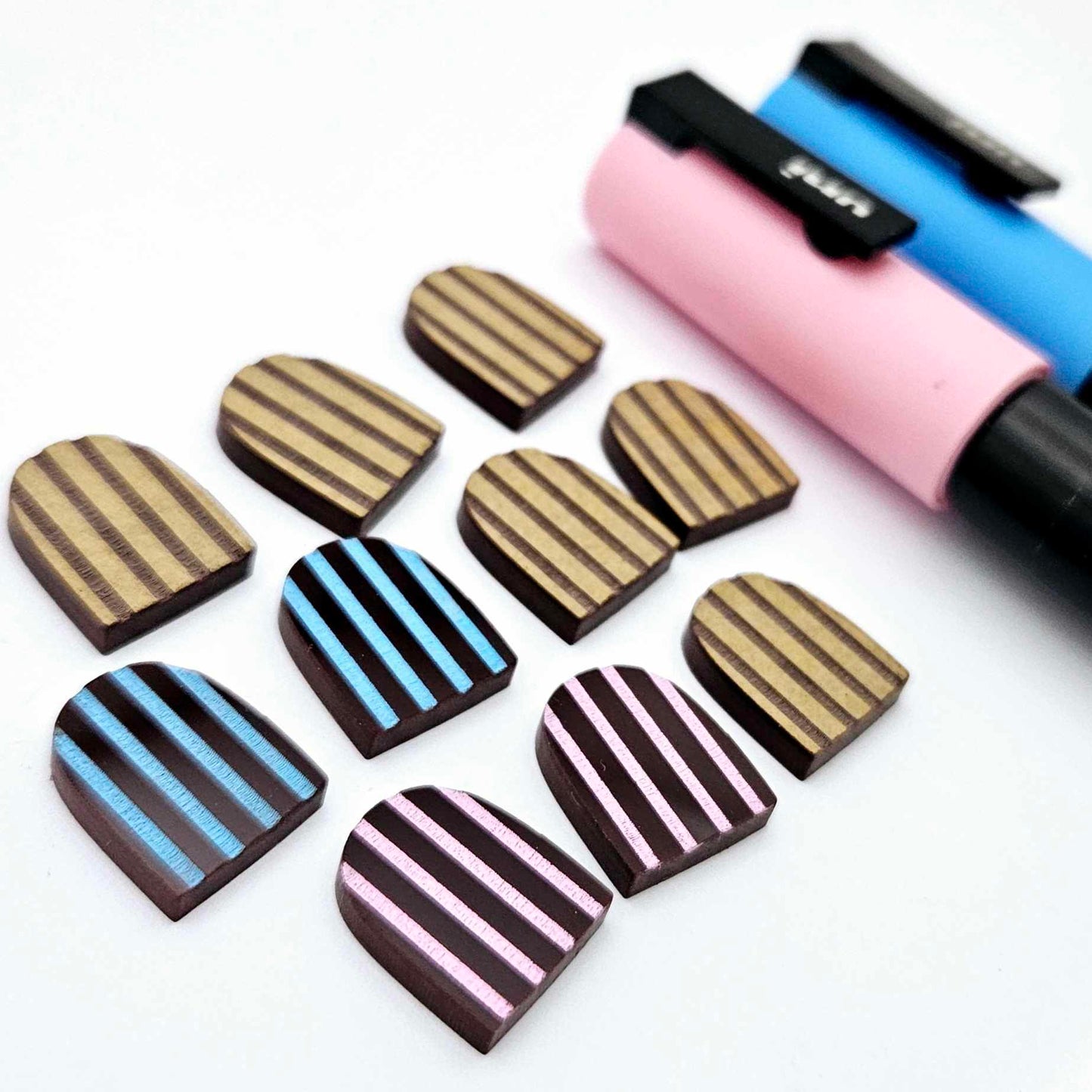 Chocolate brown striped arch stud blanks with engraved stripes, one pair paint filled light pink, one pair painted filled light blue, with additional unpainted pieces shown with protective film on