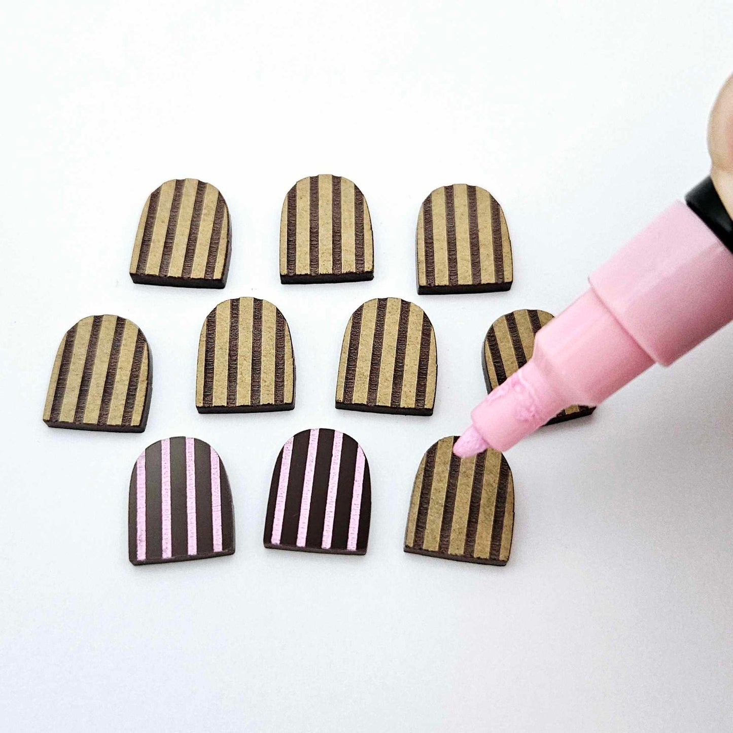 Chocolate brown striped arch stud blanks with engraved stripes, one pair paint filled light pink, with additional unpainted pieces shown with protective film on with a pink posca paint pen painting one