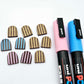 Chocolate brown striped arch stud blanks with engraved stripes, one pair paint filled light pink, one pair painted filled light blue, with additional unpainted pieces shown with protective film on