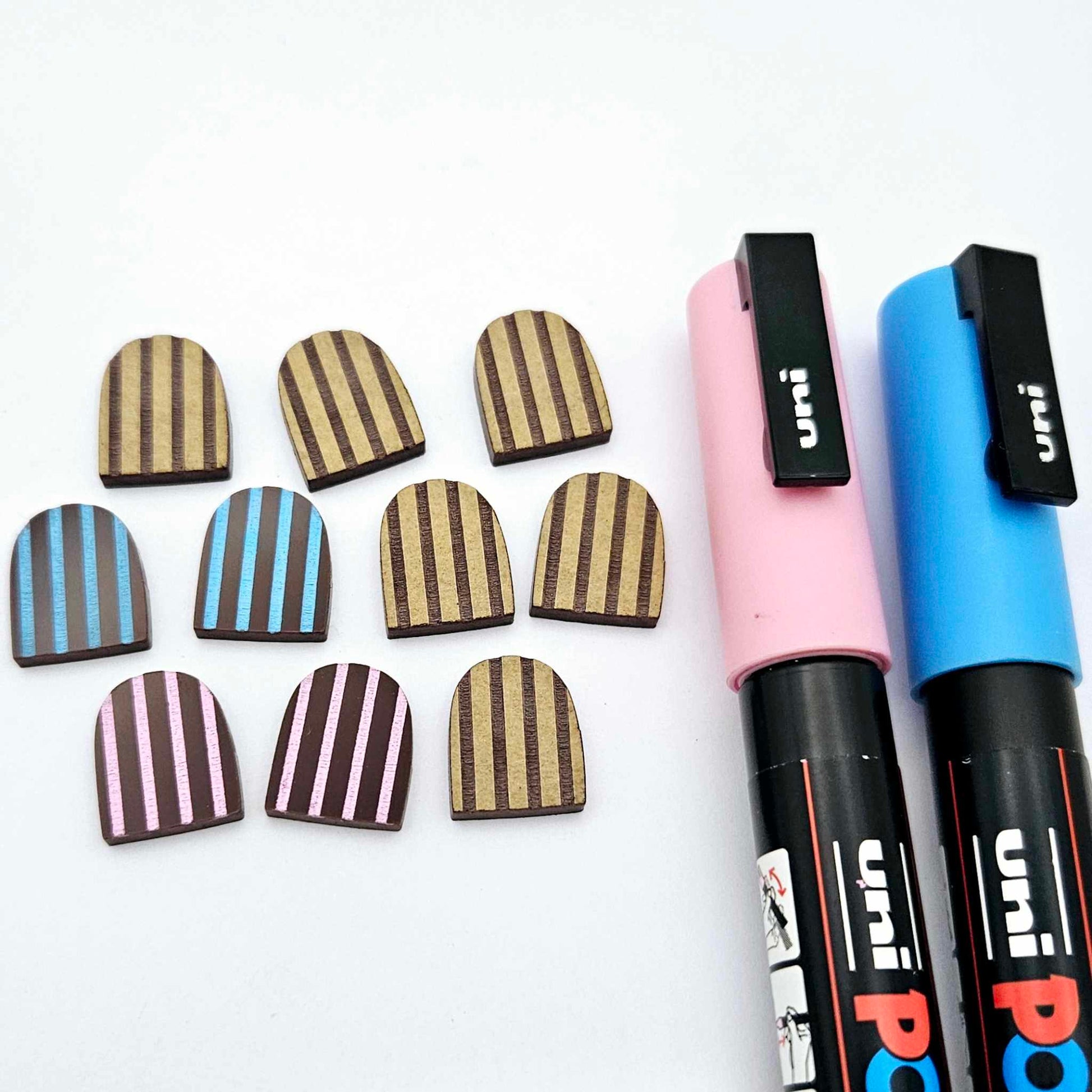 Chocolate brown striped arch stud blanks with engraved stripes, one pair paint filled light pink, one pair painted filled light blue, with additional unpainted pieces shown with protective film on