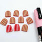 16mm red striped acrylic arch stud blanks showing paint filled example and unpainted pieces with protective film, pictured with a light pink Posca paint pen