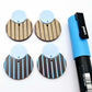 Nested circle dangle blanks showing one piece paint filled light blue and peeled, with another pair unpainted and with protective film on, pictured with a light blue Posca paint pen