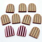 Port wine striped arch stud blanks with engraved stripes, one pair paint filled light pink and peeled, with additional unpainted pieces shown with protective film on