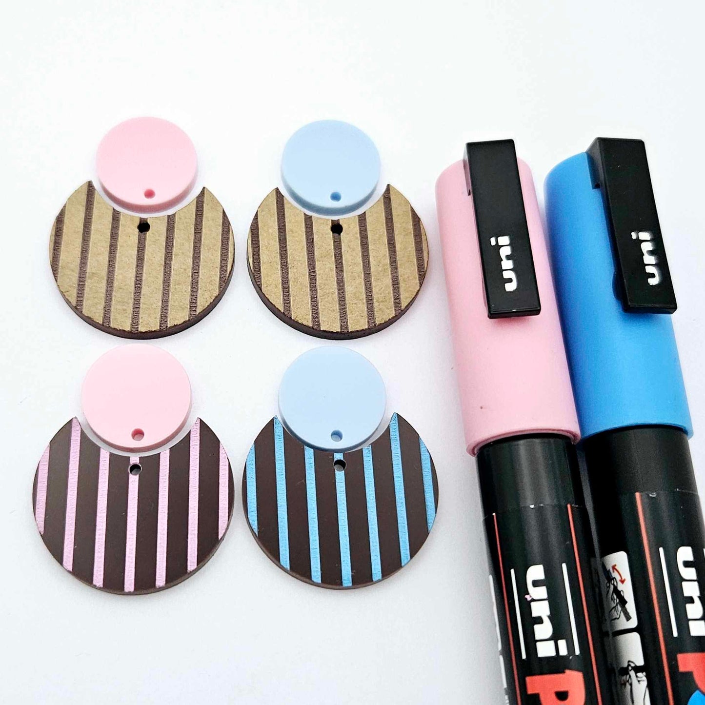 Nested circle dangle blanks showing one piece paint filled light pink and one piece paint filled light blue and peeled, with another pair unpainted and with protective film on, pictured with pink and blue Posca paint pen