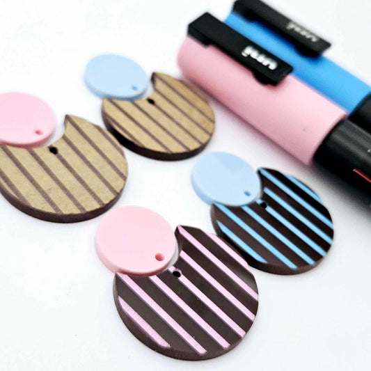 Nested circle dangle blanks showing one piece paint filled light pink and one piece paint filled light blue and peeled, with another pair unpainted and with protective film on, pictured with pink and blue Posca paint pen