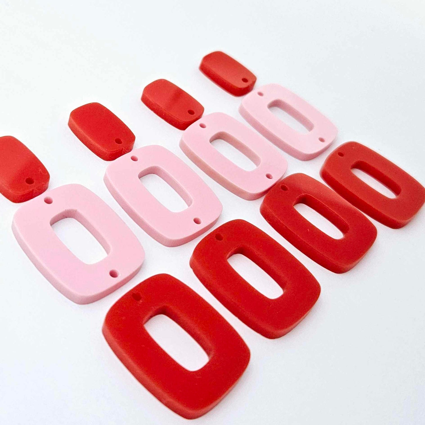 Two pairs of layered rectangle dangle blanks in red and pastel pink acrylic, unassembled pieces laid flat showing three-piece design