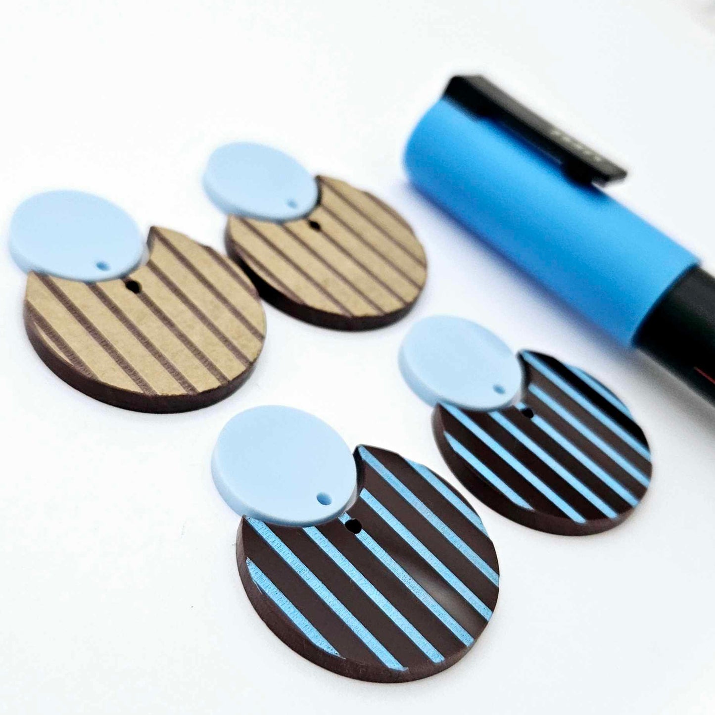 Nested circle dangle blanks showing one piece paint filled light blue and peeled, with another pair unpainted and with protective film on, pictured with a light blue Posca paint pen