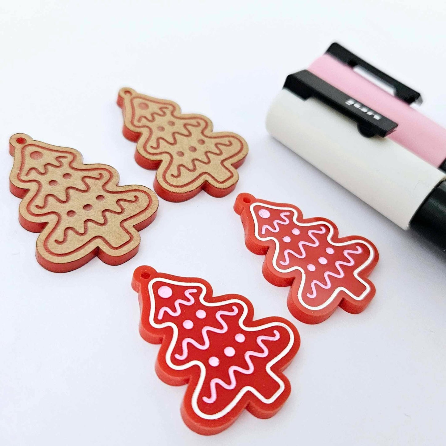 One pair of solid red cookie Christmas tree dangle earring blanks paint filled in light pink and white, alongside one unpainted pair with protective paper still attached, pictured with Posca paint pens