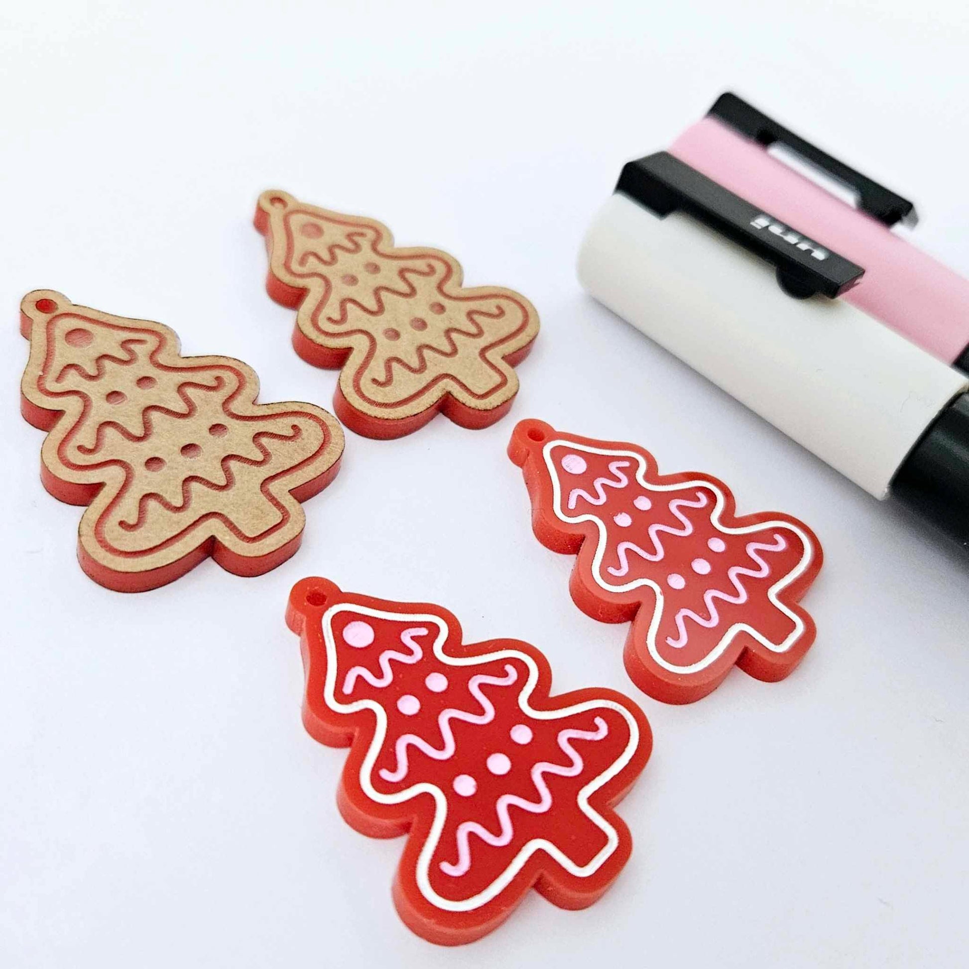 One pair of solid red cookie Christmas tree dangle earring blanks paint filled in light pink and white, alongside one unpainted pair with protective paper still attached, pictured with Posca paint pens