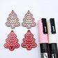 One pair of deep red marble cookie Christmas tree dangle blanks paint filled in light pink and white, alongside one unpainted pair with protective paper attached, pictured with Posca paint pens