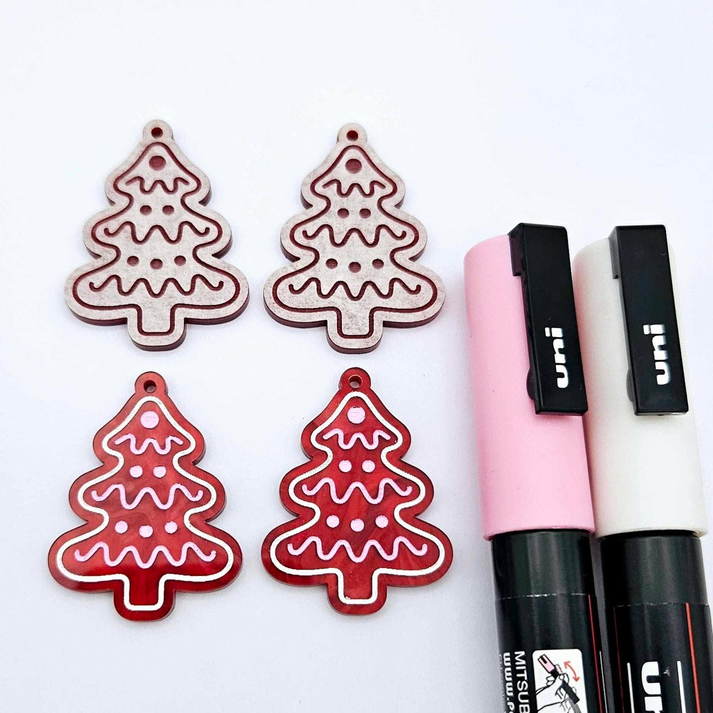 One pair of deep red marble cookie Christmas tree dangle blanks paint filled in light pink and white, alongside one unpainted pair with protective paper attached, pictured with Posca paint pens