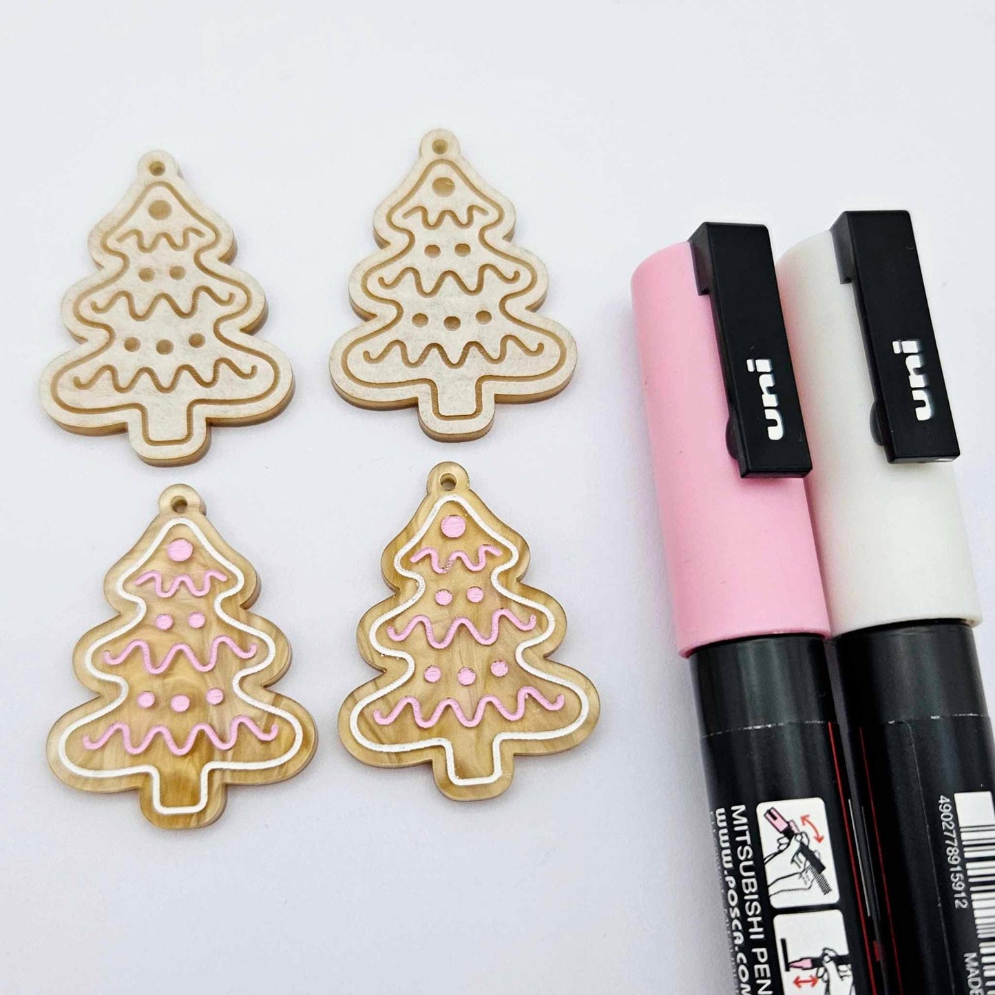 One pair of gold pearl marble cookie Christmas tree dangle earring blanks paint filled in light pink and white, alongside one unpainted pair with protective paper attached, pictured with Posca paint pens