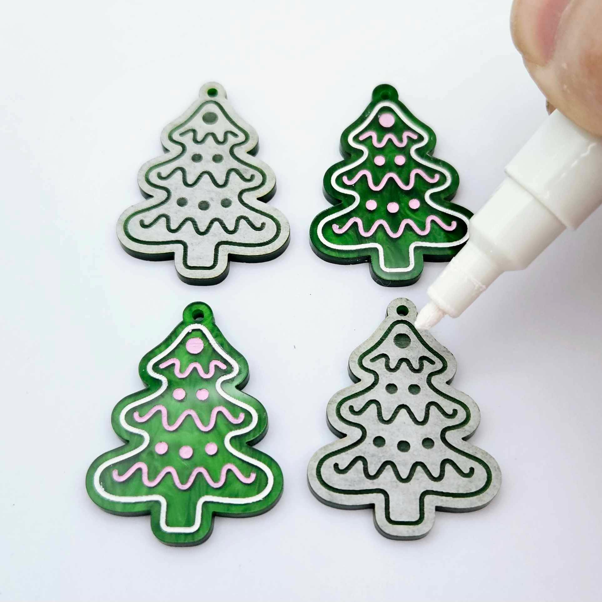 One pair of paint-filled green marble cookie Christmas tree dangle blanks beside one unpainted pair with protective paper attached, with an uncapped Posca paint pen positioned ready to paint fill the engraved details