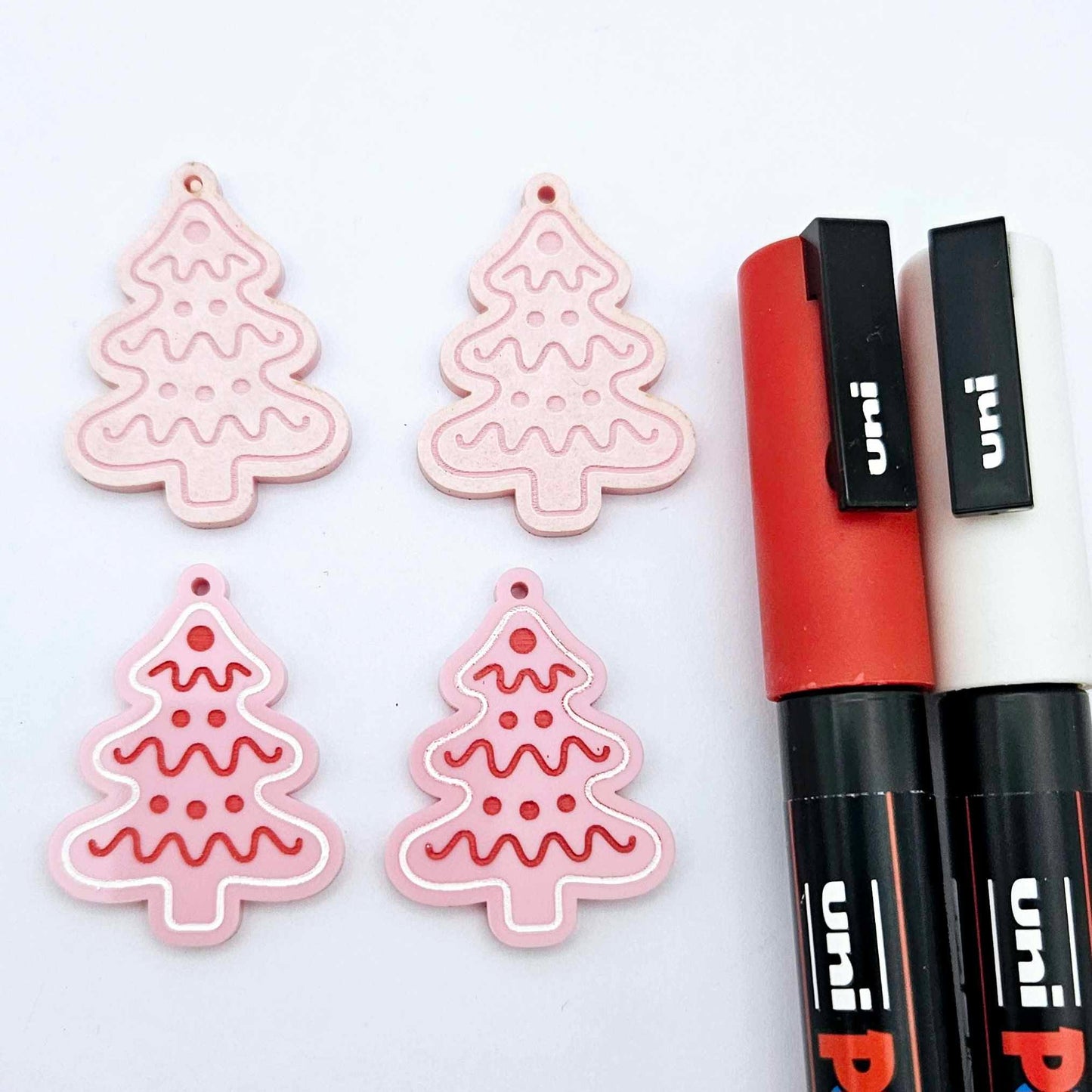 One pair of pastel pink cookie Christmas tree dangle blanks paint filled in red and white, alongside one unpainted pair with protective paper still attached, pictured with Posca paint pens