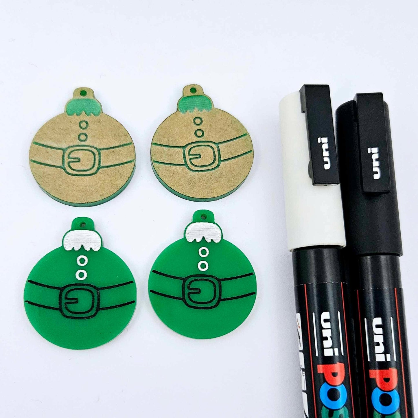 One pair of solid green Santa bauble dangle blanks paint filled in black and white, alongside one unpainted pair with protective paper attached, pictured with Posca paint pens